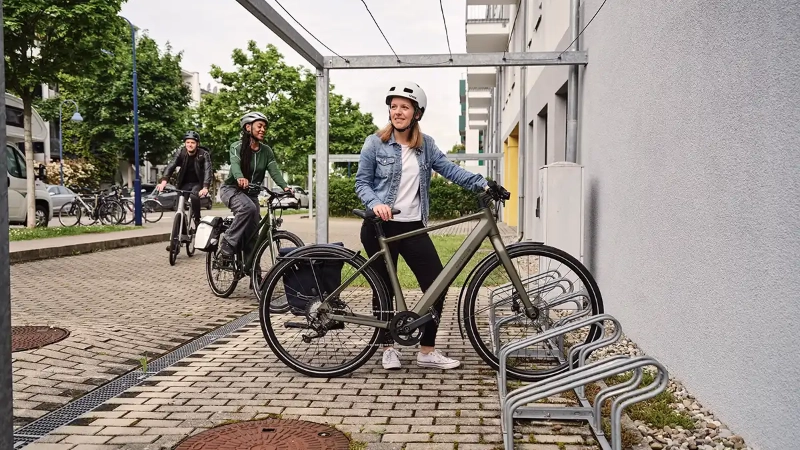Eine Frau in blauer Jeansjacke parkt ihr grünes E-Bike, eine weitere Frau und ein Mann fahren den Fahrradstellplatz an Eine Frau in blauer Jeansjacke parkt ihr grünes E-Bike, eine weitere Frau und ein Mann fahren den Fahrradstellplatz an