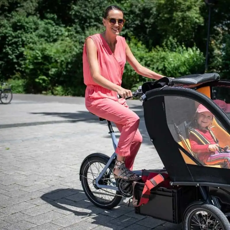 Eine Mutter fährt das Chike e-Bike in der Stadt. Ihr Kind sitzt angeschnallt darin. Beide lachen Eine Mutter fährt das Chike e-Bike in der Stadt. Ihr Kind sitzt angeschnallt darin. Beide lachen